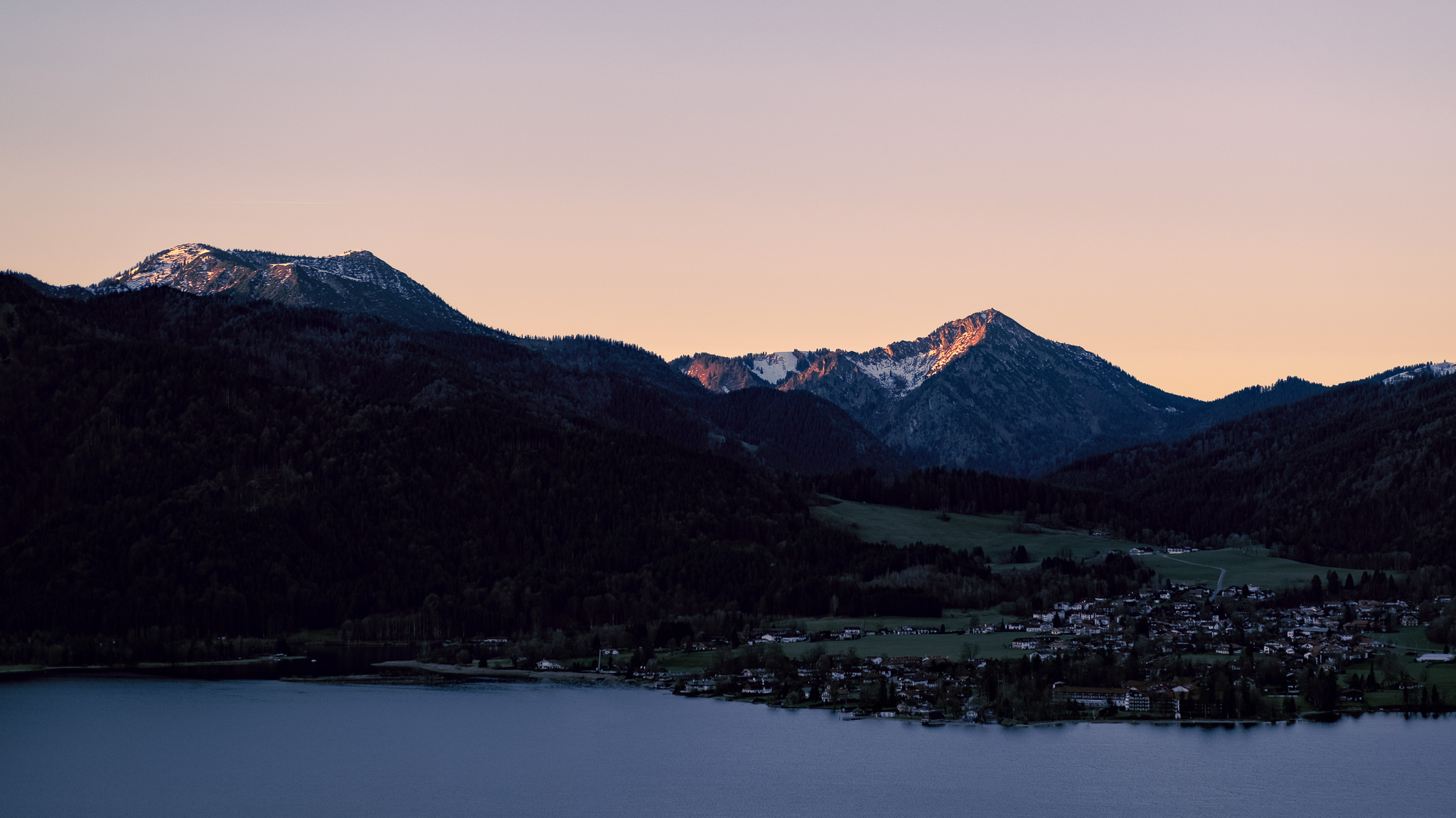 Ein Gipfel wird von rechts von der Sonne angeschienen und erstrahlt in warmen Gelbtönen, im Vordergrund ist ein See und ein daran liegendes Dorf zu sehen.